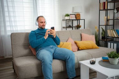 Yetişkin bir adam rahat bir koltukta otururken telefonda müzik ve sosyal medyadan keyif alıyor.