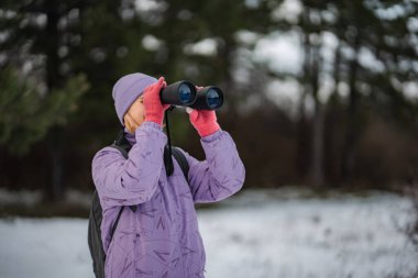 Karlı ormanda duran kadın dürbünle bakıyor, kış doğasını keşfediyor.