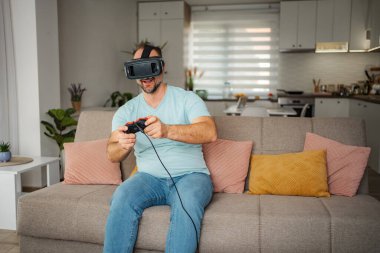 VR kulaklık takan, kumandayla kanepede oyun oynayan, boş vakitlerin tadını çıkaran bir adam.