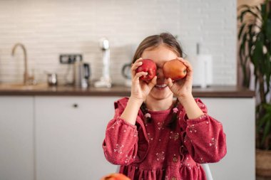 Mutlu çocuk kırmızı elmalarla gözlerini kapatıyor, parlak bir mutfakta gülümsüyor.