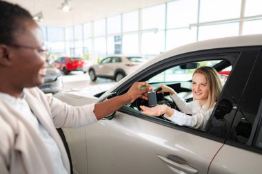 Satıcı kadın yeni arabasında oturan gülümseyen bir kadına arabanın anahtarlarını veriyor.