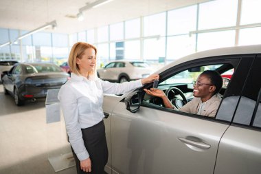 Kadın satıcı, yeni arabasında oturan gülümseyen kadın müşteriye araba anahtarı veriyor.