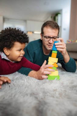 Baba ve küçük oğlu yumuşak bir halıda ahşap yapı taşlarıyla birlikte oynuyorlar.