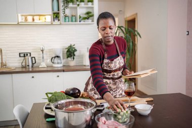 Önlüklü kadın sağlıklı yemek pişiriyor, malzemeler için bir yemek kitabına danışıyor.