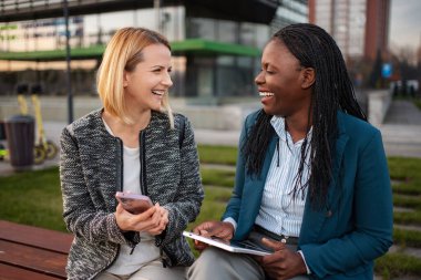 İki iş kadını bir bankta gülüyor ve sohbet ediyorlar, ellerinde telefon ve tablet var.