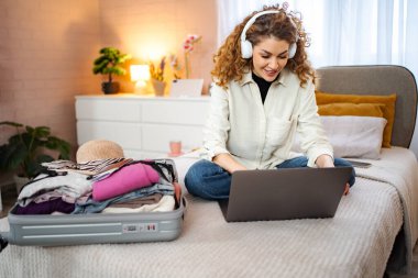 Genç yetişkin bir kadın yatak odasında giysilerini toplarken internetten gezi planlıyor.