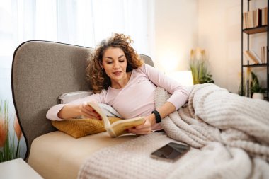 Kadın yatakta kitap okuyor, evinde rahat ve rahat bir yaşam tarzını kucaklıyor.