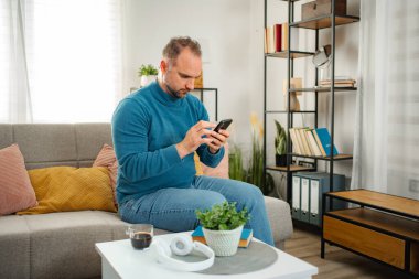 Yetişkin bir adam kanepede oturuyor, akıllı bir telefonda konsantre bir ifadeyle daktilo yazıyor.