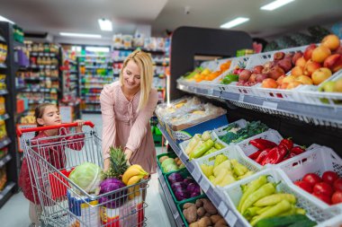 Anne ve kızı sağlıklı taze meyve ve sebzeleri alışveriş arabasına koyuyorlar.