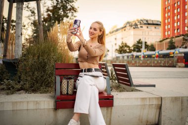 Genç yetişkin kadın gezgin Viyana 'da bir bankta otururken akıllı telefonuyla selfie çekiyor.
