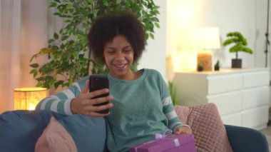 Evde kanepede otururken doğum günü hediyesini gösteren mutlu bir kadın.