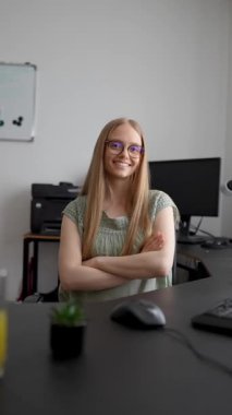 Masasında oturan genç iş kadını kollarını kavuşturmuş, gülümsüyor ve kameraya bakıyor.