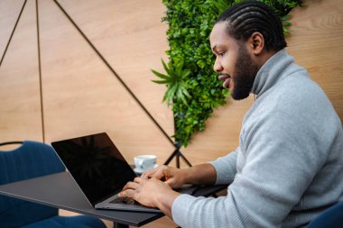 Genç yetişkin bir adam, modern bir kafede masada otururken dizüstü bilgisayarla telgraf çekiyor.