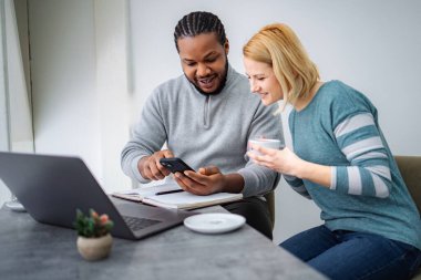 Mutlu çift gülümsüyor ve masada çalışırken bir akıllı telefon ekranını paylaşıyor.