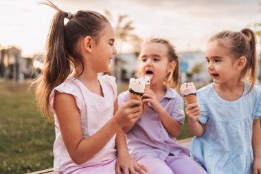 Üç küçük kız bir bankta oturuyor, yaz günü dondurma külahlarının tadını çıkarıyorlar.