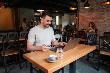 Genç işadamı bir kafede çalışırken dizüstü bilgisayar ve akıllı telefon kullanıyor.