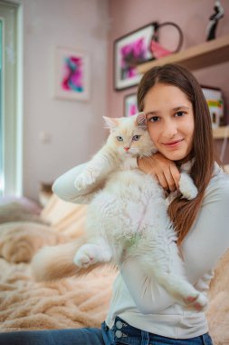 Mavi gözlü, tüylü beyaz kediyi tutan kız gülümsüyor.