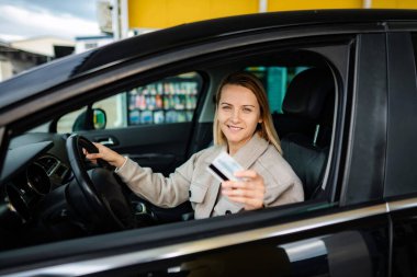 Arabanın camından kredi kartı tutan bir kadın, benzin istasyonunda gülümsüyor.