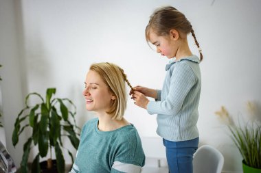 Küçük kız annesinin saçlarını örüyor, özen ve beraberlik bağı kuruyor.