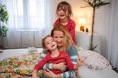 Anne, evdeki yatakta iki mutlu kızıyla sarılıp oynuyor.