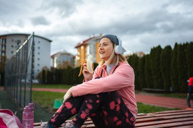 Spor giyim bölümünde müzik dinleyen, muz yiyen, antrenmandan sonra dinlenen bir kadın.