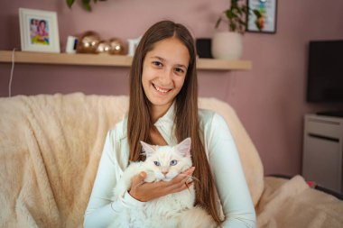 Genç kız kedisini rahat bir kanepede tutarken kameraya gülümsüyor.