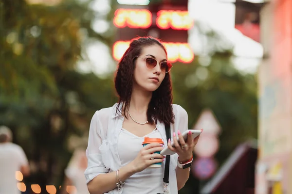 A woman in sunglasses walks with coffee on the street.