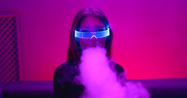 Mulher jovem voa vape em luz de néon em óculos cibernéticos. Há um efeito de grão, barulho. — Vídeo de Stock