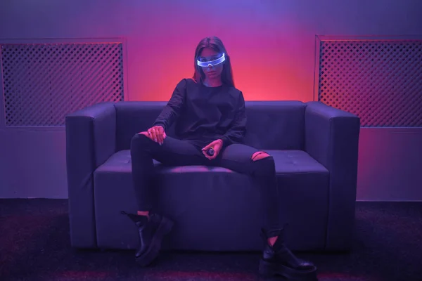 Cyberpunk γυναίκα κάθεται στον καναπέ με γυαλιά νέον. Η φωτογραφία έχει ως αποτέλεσμα σιωπή, σιτάρι. — Φωτογραφία Αρχείου