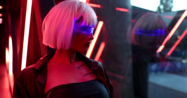 The blonde in neon glasses sits in a futuristic room and looks out the window. — Stock Video