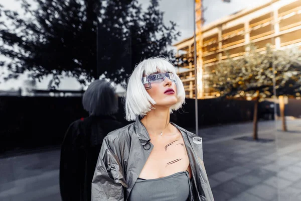 Stylish blonde in futuristic clothes and glasses on a mirror background. — Stock Photo, Image