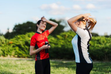 Gündüz vakti parkta koştuktan sonra iki kadın esneyip gülümsüyor. Su şişeleri var..