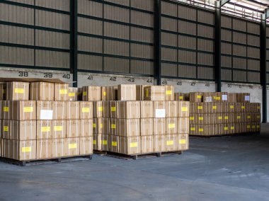 Perak, Malezya - 5 Ağustos 2022: Ahşap paletler üzerine istiflenmiş kahverengi karton karton kutular, koridor numarasına göre düzenlenmiş, lojistik, dağıtım ve depolama için geniş ve boş bir sanayi deposunda hazır..