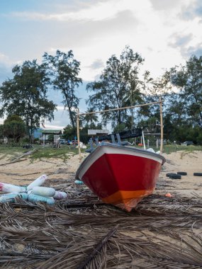 Geleneksel kırmızı ve turuncu fiberglas balıkçı teknesinin alçak açılı çekimi Dungun, Terengganu 'daki kumlu bir sahilde çekildi. Kurumuş palmiye yaprakları ve şamandıralarıyla çevrili..