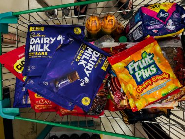 Perak, Malezya - 17 Aralık 2025: Bir süpermarkette içinde Cadbury Süt Mini barları, Fruit Plus Chewy Candy ve Libresse pedleri bulunan yeşil metal alışveriş arabasının yukarıdan görünüşü.