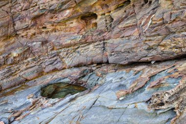Renkli mineral katmanları, yıpranmış taş dokusu ve küçük bir gelgit havuzu gösteren jeolojik tortul kaya oluşumunun yakın çekimi..