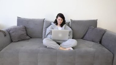 Evde dizüstü bilgisayarda çalışan bir kadın, koçun üstünde oturuyor.