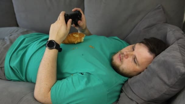 Hombre jugando videojuegos comer croissant acostado en el entrenador — Vídeo de stock