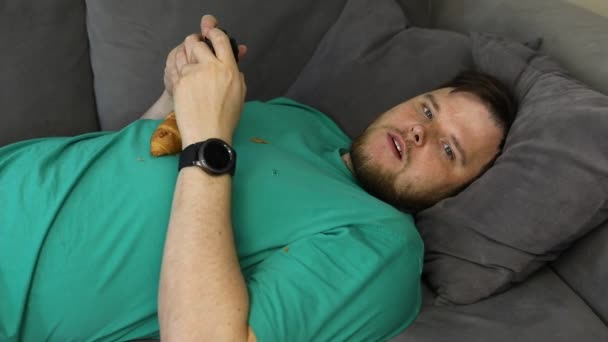 Hombre jugando videojuegos comer croissant acostado en el entrenador — Vídeo de stock