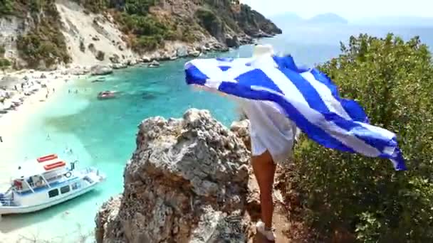 Жінка з прапором Греції дивиться з вершини гори на морському пляжі — стокове відео