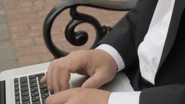 Empresario escribiendo en un portátil y sentado en un banco en la calle — Vídeos de Stock