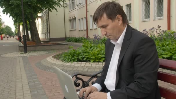 Businessman typing on a laptop and sitting on a bench in the street — Stock Video