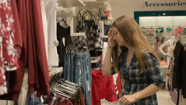 Joven chica bonita está eligiendo la ropa en la tienda en el centro comercial — Vídeos de Stock