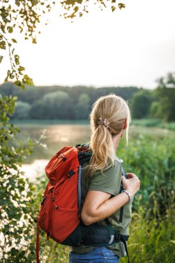 Kırmızı sırt çantalı yürüyüşçü kadın doğada göle bakıyor. Açık hava macerası ve keşfi