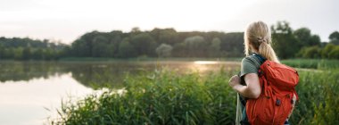 Kırmızı sırt çantalı kadın yürüyüşçü doğa yürüyüşü yaparken göl veya göl manzarasının keyfini çıkarıyor. Açık hava macerası