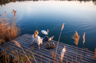 Huzurlu göl manzarası. Kuğu yakınlarda yüzerken bir kadın iskelede dinleniyor. Doğayla bağlantı ve uyum. Nadir ve barışçıl karşılaşma