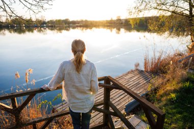 Açık havada tefekkür ve farkındalık. Tahta terasta duran kadın, sakin göle bakıyor ve doğada sessiz bir anın ve günbatımının tadını çıkarıyor.