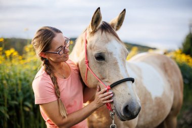 Gülümseyen kadın çayırda atıyla bağ kuruyor. Altın akşam ışığında güven ve duygusal bir bağ gösteriyor.