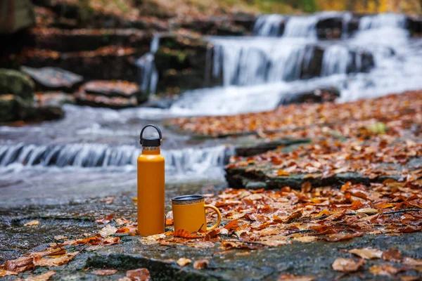 Sonbahar ormanındaki şelalede termos ve seyahat kupası. Yürüyüş ekipmanları. Sıcak içeceği bütün gün saklamak için izole edilmiş şişe.