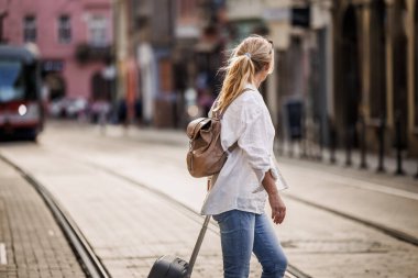 Bavullu bir turist şehirde tramvay raylarıyla caddeyi geçiyor. Seyahat ve tatil yaşam tarzı